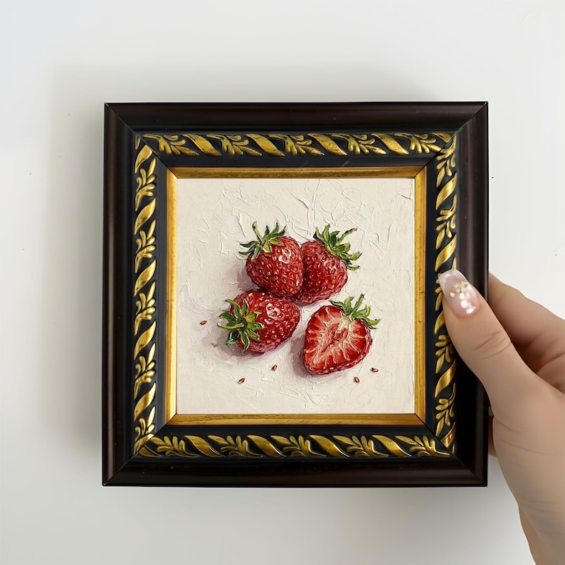 May include: A framed painting of strawberries. The artwork features four ripe, red strawberries with green leaves against a textured white background. The frame is dark brown with gold accents. The painting is held by a hand.