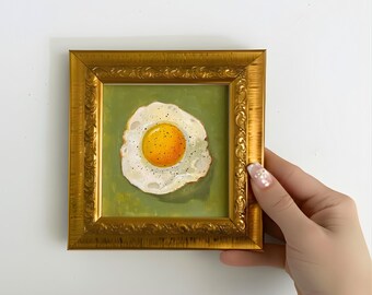 May include: A framed painting of a fried egg. The egg has a bright yellow yolk with pepper and a white egg white, set against a green background. The frame is gold with ornate detailing.