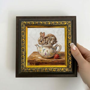 May include: An oil painting in a dark brown and gold ornate frame depicting a small brown rabbit sitting on a white floral teapot. The teapot rests on a wooden surface next to a single red strawberry, all set against a light, textured background.