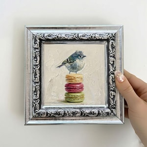 May include: An oil painting in a silver ornate frame depicting a small blue and grey bird perched atop a vertical stack of three colourful macarons. The macarons are coloured light yellow, magenta, and light green, set against a textured cream background.