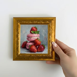 May include: A framed painting of pink macarons and fresh strawberries. The macarons are stacked, with strawberries scattered around them. The painting is in a gold ornate frame, held by a hand.