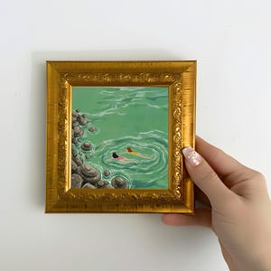Könnte beinhalten: Ein gerahmtes Gemälde zeigt zwei Figuren, die in grünem Wasser schwimmen. Das Kunstwerk ist in einem goldenen, verzierten Rahmen gefasst. Das Wasser hat ein kreisförmiges Muster, und Felsen sind am Rand des Wassers zu sehen.