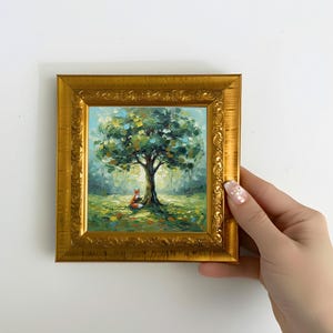 Puede incluir: Pintura al óleo enmarcada que representa una serena escena de bosque. Un marco dorado rodea la obra de arte, mostrando un gran árbol con exuberante follaje verde. Un pequeño zorro está sentado en la base del árbol. El estilo es suave e impresionista.