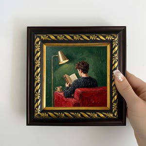 以下が含まれることがあります： 金色のランプの下、赤いアームチェアで本を読む人物を描いた額入りの絵画。アートワークは、金色のアクセントが付いた濃い茶色のフレームが特徴です。背景はテクスチャ加工された緑色で、小さなテーブルの上にコーヒーカップが置かれています。