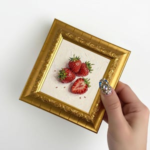 May include: A small, square, gold-framed painting featuring a still life of strawberries. The painting depicts four ripe red strawberries, one sliced open, on a textured white background. The frame has an ornate, detailed design.