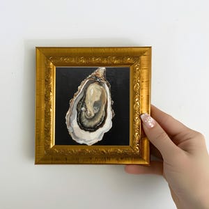 May include: A framed painting of an oyster on a black background. The oyster is open, revealing a pearlescent interior with dark gray and white details. The frame is ornate gold with a square shape.