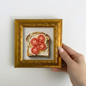 以下が含まれることがあります： バターとスライストマトを添えたトーストのスライスを描いた額入りの絵画。アートワークは金色の装飾フレームに入っています。絵は肌の明るい人が持っています。