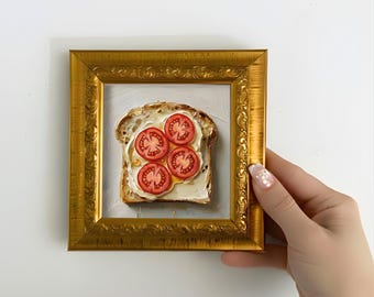 May include: A framed painting of a slice of toast with butter and sliced tomatoes. The painting is in a gold ornate frame. The artwork is held by a person with a light skin tone.
