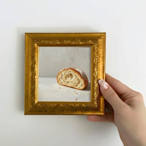 Puede incluir: Pintura enmarcada de una rebanada de pan. El pan tiene una corteza dorada y un interior suave y texturizado. La pintura está enmarcada en un marco dorado ornamentado.