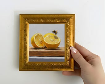 May include: A framed still life painting featuring halved lemons and a bumblebee. The lemons are yellow with a light brown cutting board. The frame is gold with ornate detailing. The painting is held by a hand.