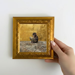 May include: A framed artwork featuring a small monkey reading a book. The artwork is set against a gold background and a textured foreground. The frame is ornate and gold-colored, adding a touch of elegance to the piece.