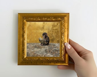 May include: A framed artwork featuring a small monkey reading a book. The artwork is set against a gold background and a textured foreground. The frame is ornate and gold-colored, adding a touch of elegance to the piece.