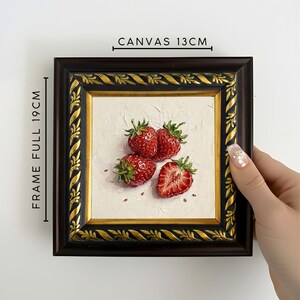 May include: A framed painting of strawberries. The canvas measures 13cm, and the full frame is 19cm. The painting features four red strawberries with green leaves on a white background. The frame is dark brown with gold accents.