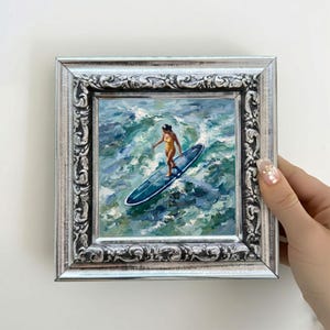May include: An oil painting in a decorative silver frame depicting a person in a yellow swimsuit standing on a blue surfboard amidst churning turquoise and white ocean waves. The artwork features visible brushstrokes and a textured, painterly style.