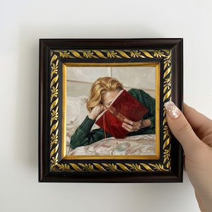 Puede incluir: Pintura enmarcada que representa a una persona leyendo un libro en la cama. La persona lleva un suéter verde y sostiene un libro rojo. El marco marrón oscuro tiene detalles dorados.