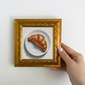 Puede incluir: Pequeña pintura enmarcada de un cruasán sobre un plato blanco. El cruasán es de color marrón dorado y se encuentra sobre un plato blanco con una sombra sutil. El marco es dorado y ornamentado, y la pintura es sostenida por una mano.