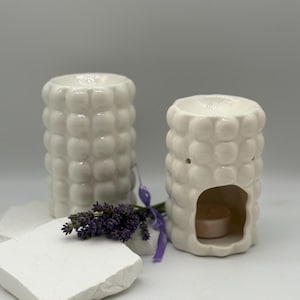 Könnte beinhalten: Zwei weiße Keramik-Duftzerstäuber mit einem strukturierten, blasenartigen Design. Ein Diffusor hat eine Öffnung für ein Teelicht. Ein Zweig lila Lavendel und ein weißer Stein befinden sich im Vordergrund.