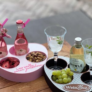 Könnte beinhalten: Zwei ovale Getränke- und Snackhalter mit gestreiften Oberflächen. Ein rosa Halter enthält zwei Spritz-Flaschen sowie Schalen mit getrockneten Erdbeeren und Nüssen. Ein schwarzer Halter enthält eine Bowle-To-Go-Flasche, grüne Trauben und zwei Gläser.