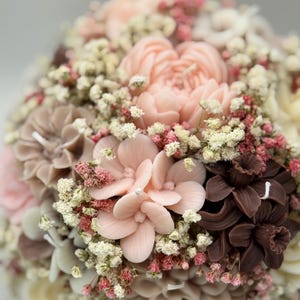 Könnte beinhalten: Nahaufnahme eines Blumenarrangements mit verschiedenen blütenförmigen Kerzen in Rosa-, Braun- und Cremetönen. Das Arrangement umfasst kleine weiße und rosa Akzentblüten, die ein strukturiertes, geschichtetes Aussehen erzeugen. Jede Kerze hat einen Docht.