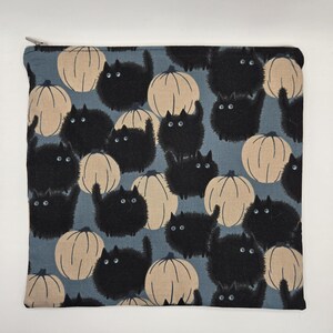 May include: A square, zippered pouch featuring a repeating pattern of black cats and beige pumpkins on a blue background. The cats have wide eyes and fluffy fur. The pumpkins have vertical lines. The pouch is likely made of fabric.