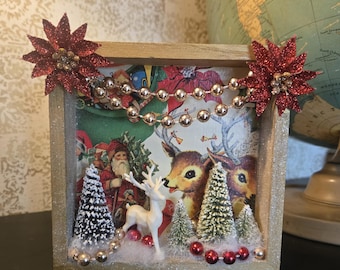 Puede incluir: Un marco cuadrado de madera tipo caja con una escena navideña que incluye una ilustración vintage de Santa Claus y renos. La decoración presenta árboles miniatura, un ciervo de plástico blanco, guirnaldas de cuentas doradas y flores de pascua rojas con purpurina.