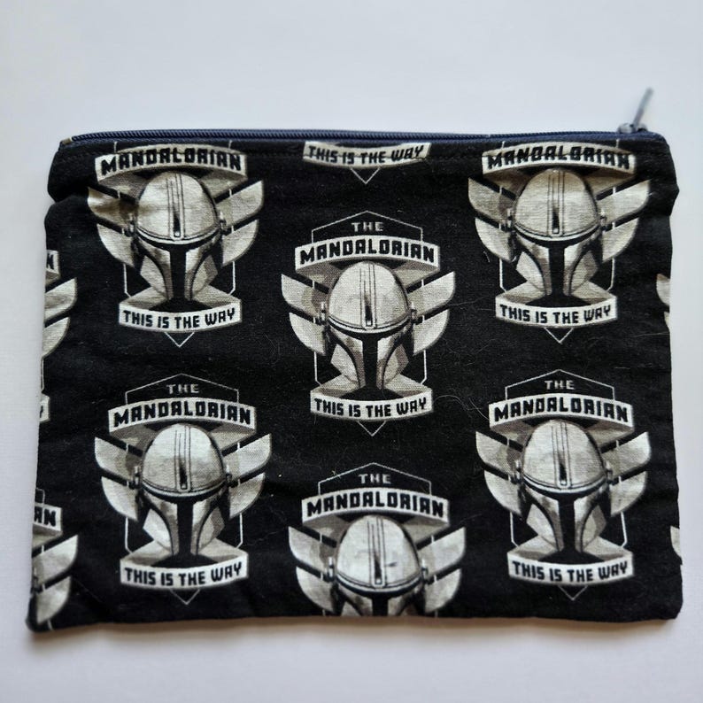 May include: Black zippered pouch featuring a repeating pattern of The Mandalorian helmet design. The design includes the words "The Mandalorian" and "This is the Way" in white. The pouch has a navy blue zipper and trim.
