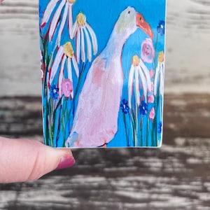 Puede incluir: Un ganso blanco con un pico rosa se encuentra en un campo de margaritas blancas y flores azules sobre un fondo azul. El texto "DO GREAT THINGS" est&aacute; en la parte superior de la imagen.