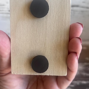 Puede incluir: Un bloque de madera marr&oacute;n claro con dos imanes redondos negros unidos a la parte posterior.
