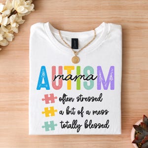 May include: White t-shirt with the text "AUTISM mama" in rainbow colors. Below the text are three puzzle pieces with the text "often stressed", "a bit of a mess", and "totally blessed".