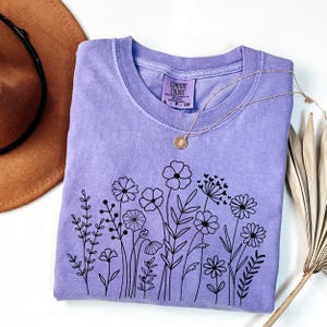 May include: A lavender t-shirt with a black floral design. The shirt has a crew neck and short sleeves. The text "COMFORT COLORS" is on the tag.