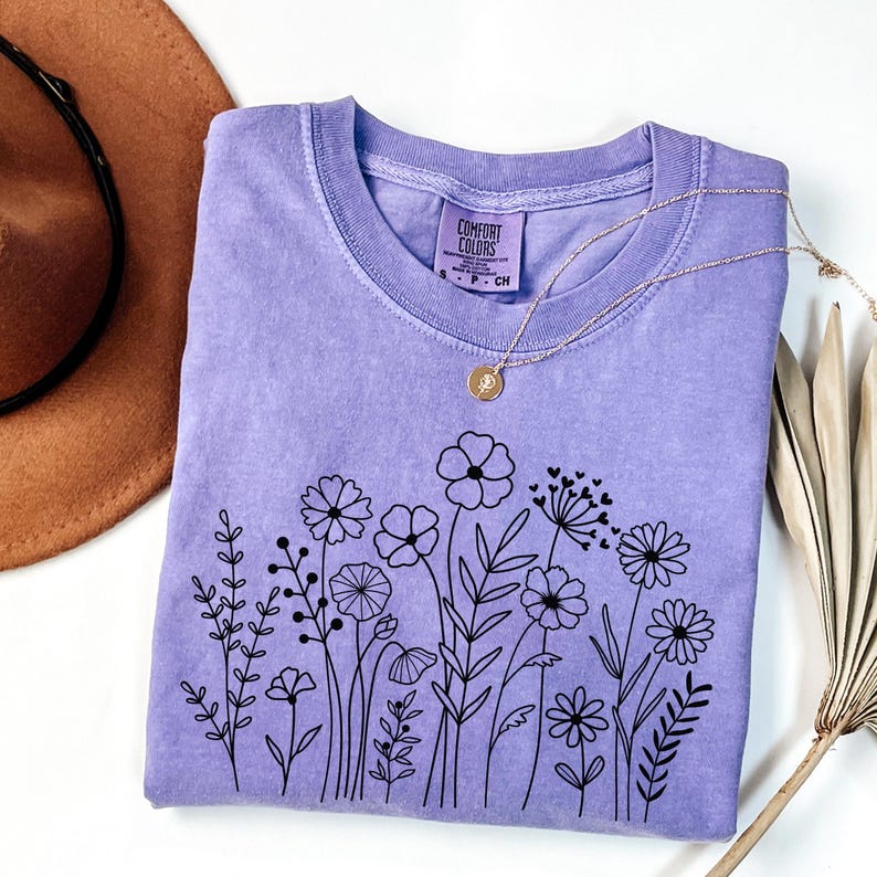 May include: A lavender t-shirt with a black floral design. The shirt has a crew neck and short sleeves. The text "COMFORT COLORS" is on the tag.