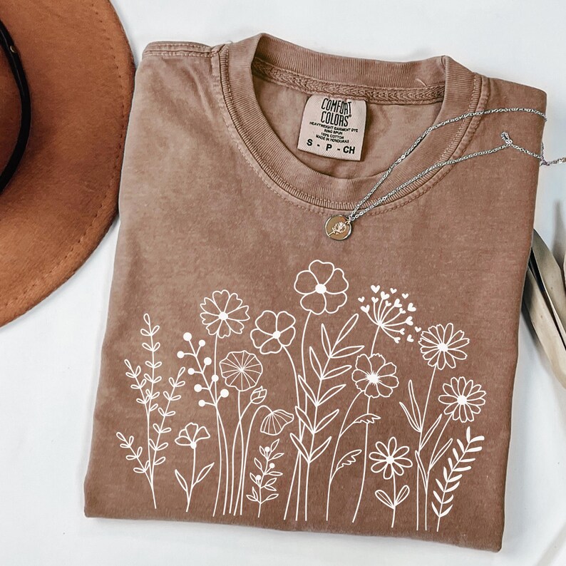 May include: A brown t-shirt with a white floral design. The shirt has a crew neck and short sleeves. The text "COMFORT COLORS" is visible on the tag.