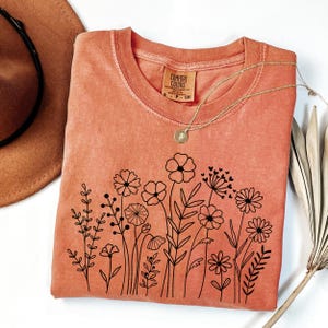 May include: A burnt orange t-shirt with a black floral design. The shirt has a crew neck and is made of a soft, comfortable fabric. The design features a variety of flowers and leaves, all outlined in black. The shirt is perfect for everyday wear.