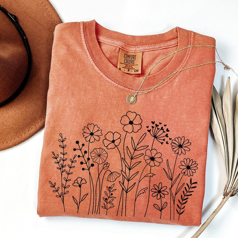 May include: A burnt orange t-shirt with a black floral design. The shirt has a crew neck and is made of a soft, comfortable fabric. The design features a variety of flowers and leaves, all outlined in black. The shirt is perfect for everyday wear.