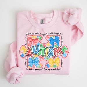 May include: Pink sweatshirt with a colorful puzzle piece design that says "AUTISM". The puzzle piece is made up of bows and the text around the puzzle piece says "I would not change you for the world, but I would change the world for you".
