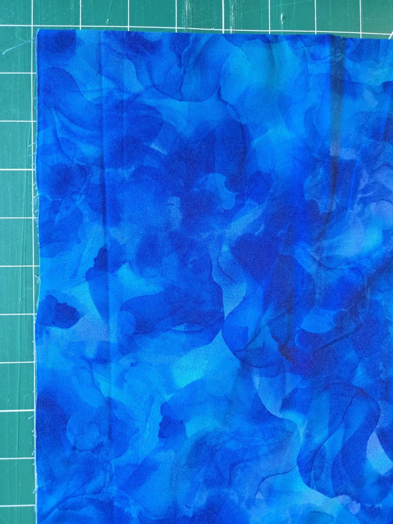 May include: Close-up of a piece of fabric with a vibrant blue abstract pattern. The fabric features shades of blue, creating a watercolor effect. The fabric is on a green cutting mat.