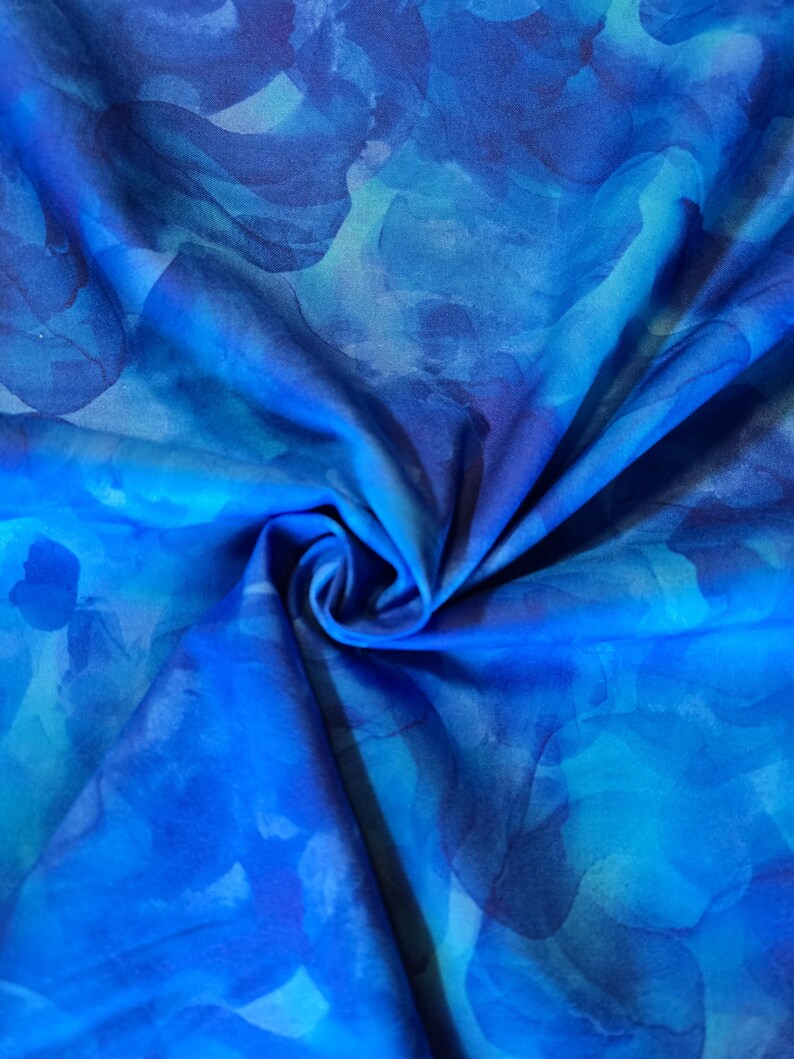 May include: A close-up of a fabric with a watercolor-like design. The fabric features shades of blue, from light turquoise to deep indigo, creating a flowing, abstract pattern. The fabric is gathered, showing folds and shadows.