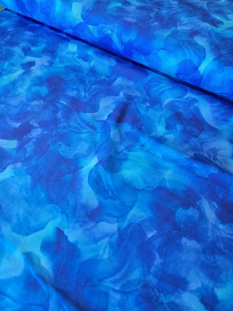 May include: A bolt of fabric with a watercolor-style floral pattern in shades of blue. The design features overlapping, abstract shapes in varying tones, creating a flowing, artistic effect. The fabric appears to be lightweight and suitable for apparel or home decor.