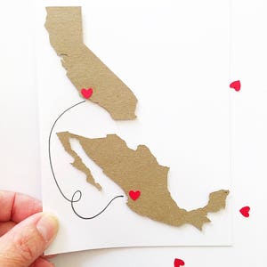 May include: A handmade card featuring a map of California and Mexico, connected by a black line. Red heart-shaped embellishments mark locations on the map. The card is held against a white background, with scattered red hearts.
