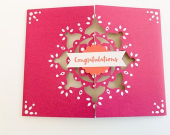 May include: A magenta-colored greeting card with a laser-cut floral design. The card features the word "Congratulations" on a banner. The card is open, revealing a beige interior.