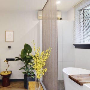 Puede incluir: Una bañera blanca con una bandeja de madera en el lateral, una cortina marrón colgando en el fondo y una planta en maceta con flores amarillas en un jarrón sobre una mesa de madera.
