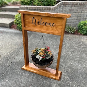 May include: A wooden welcome sign with a hanging basket of flowers. The sign is made of stained wood and features the word "Welcome" in a cursive font. The hanging basket contains a variety of colourful flowers and greenery.