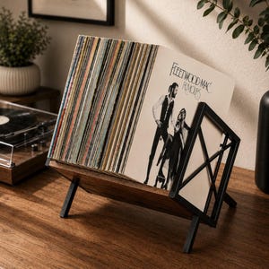 Może przedstawiać: Stojak na płyty winylowe z czarnego metalu i ciemnego drewna, zawierający kolekcję płyt, w tym album Rumours zespołu Fleetwood Mac. Stojak posiada nachyloną podstawę z czterema nogami oraz geometryczny panel boczny w kształcie litery X.