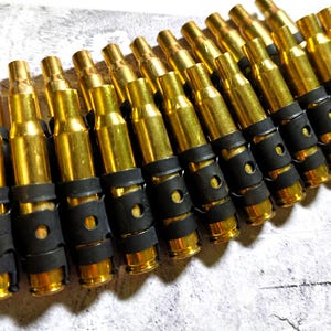 Puede incluir: Primer plano de una bandolera de casquillos de bala de latón unidos con conectores de goma negra. Los casquillos son de color dorado brillante y los conectores de goma son negros. La bandolera está en ángulo diagonalmente en el encuadre.