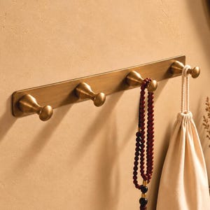 Op de afbeelding: Een wandkapstok bestaande uit een lange, rechthoekige plaat van geborsteld messing met vier bijpassende afgeronde haken. Aan de twee rechterhaken hangen een donkerrode gebedsketting en een crèmekleurig stoffen zakje met trekkoord.
