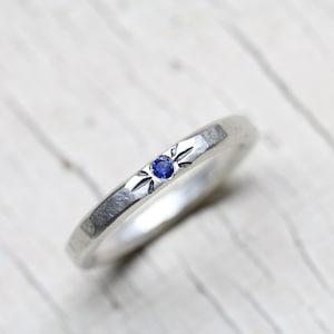 May include: A silver band ring with a small blue sapphire gemstone set in a starburst design.