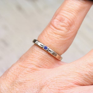 May include: A silver ring with a small blue gemstone set in the center. The ring has a simple design with a textured band.