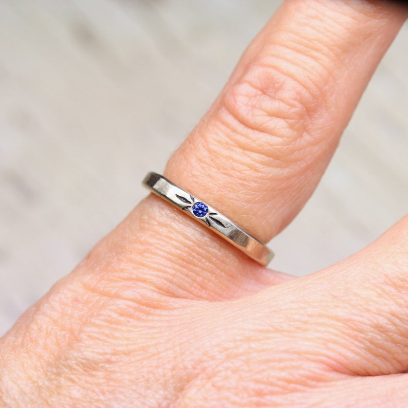 May include: A silver ring with a small blue gemstone set in the center. The ring has a simple design with a textured band.