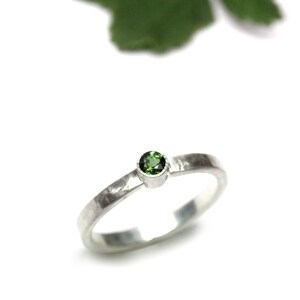 May include: A silver ring with a round, vibrant green gemstone. The band has a textured, hammered appearance. The ring is set against a white background, with a green leaf in the upper corner.