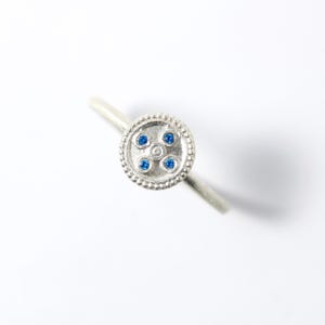 May include: A silver ring with a circular design featuring four blue gemstones and a central accent stone. The ring's band is thin and smooth, and the circular design is surrounded by a beaded border. The ring is set against a white background.
